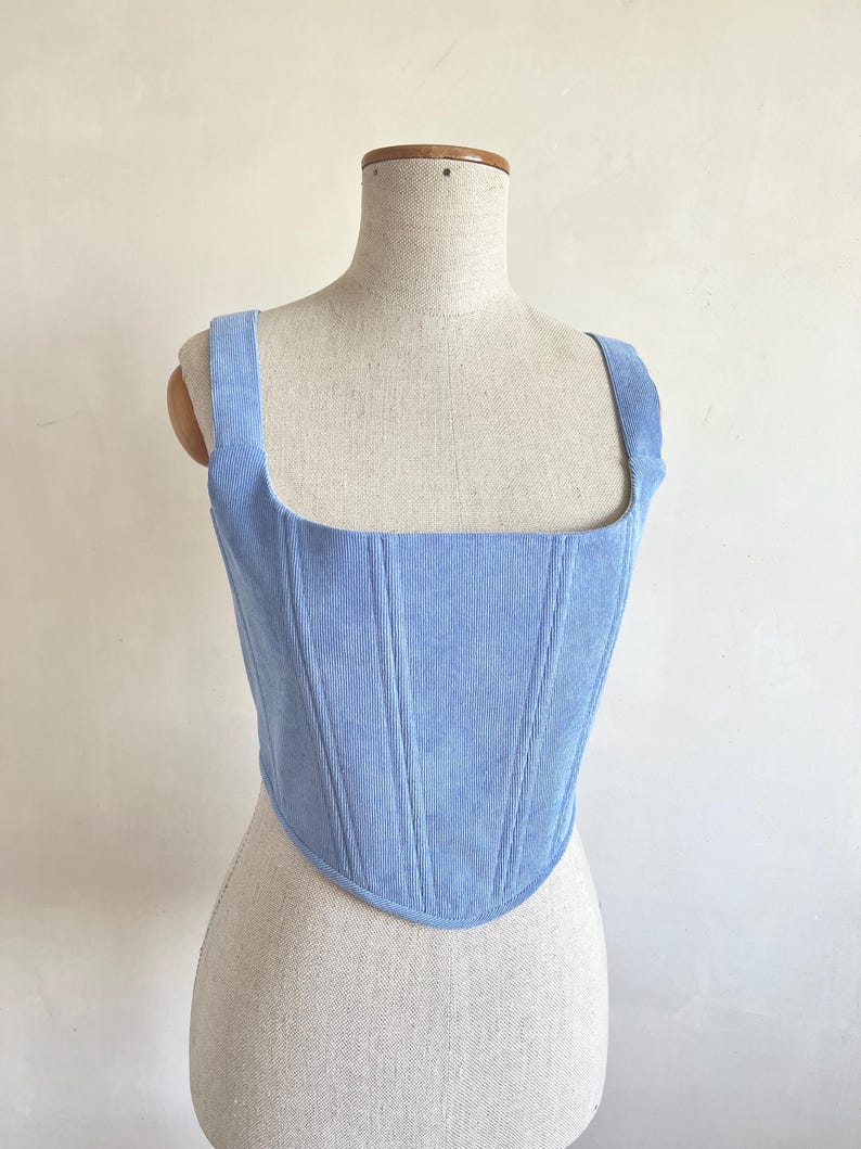 May include: A pale blue corduroy corset top with a square neckline and broad straps. The bodice has vertical seams and a curved hem. The garment is displayed on a mannequin.