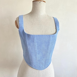 May include: A light blue corset-style top made of corduroy fabric. The garment features a square neckline, wide shoulder straps, and vertical seam detailing that creates a structured, fitted silhouette with a curved hemline at the waist.