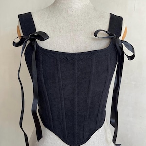 May include: A black corset-style top featuring a textured corduroy fabric, a square neckline with delicate scalloped lace trim, and wide shoulder straps adorned with long, tied black satin ribbons on each side, creating a structured silhouette.