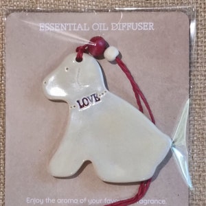 May include: White ceramic dog-shaped essential oil diffuser with the word "LOVE" on it. The diffuser is hanging from a red string with a red bead.