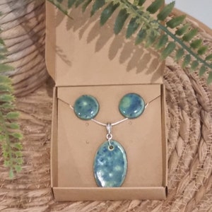 May include: A set of green and blue ceramic earrings and a pendant necklace in a brown box. The earrings are round and the pendant is oval shaped.