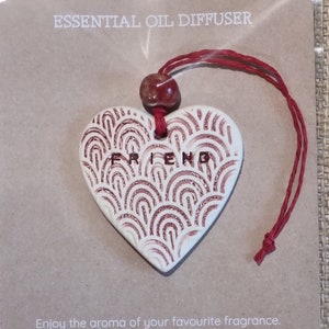 May include: White ceramic heart-shaped essential oil diffuser with a red and white pattern. The diffuser has the word "FRIEND" written on it and is hanging from a red string.