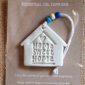 May include: White ceramic home-shaped essential oil diffuser with the text "Home Sweet Home" and a heart. The diffuser hangs from a white string with blue and white beads.