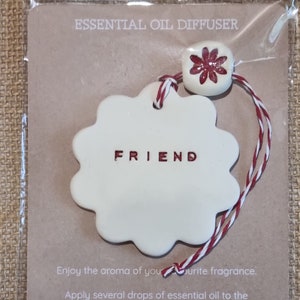 May include: White ceramic diffuser with the word "FRIEND" in red lettering. The diffuser has a red flower charm and a red and white string.