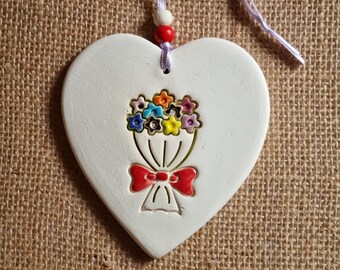 May include: White ceramic heart-shaped ornament with a red bow and a bouquet of colourful flowers. The ornament has a hole at the top for hanging.