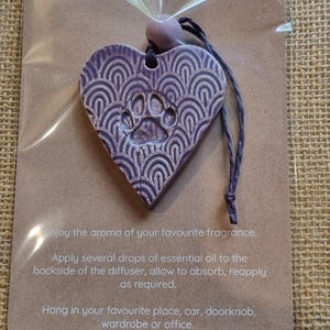 May include: A lavender heart-shaped essential oil diffuser with a paw print design and a decorative pattern. It includes a string for hanging and the text "ESSENTIAL OIL DIFFUSER".