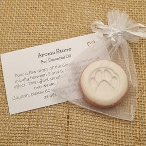 May include: Aroma stone for essential oil, with a white paw print design. The round stone has a red border, presented in a sheer white organza bag with a tag that reads "Aroma Stone For Essential Oil".