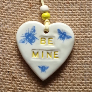 May include: White ceramic heart-shaped ornament with a yellow string and beads. The ornament has three blue bee illustrations and the text "BE MINE" in yellow.