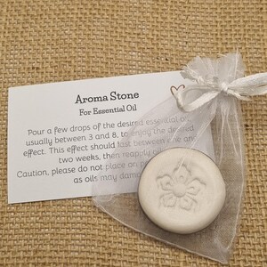 May include: Aroma Stone for Essential Oil. A white, circular stone with a floral pattern, in a sheer white bag tied with a ribbon. A card with instructions is visible. The stone is designed for use with essential oils.