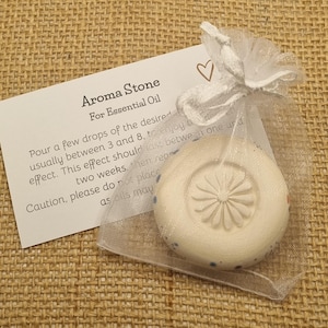 May include: Aroma stone for essential oil, packaged in a sheer white bag. The stone is round, off-white, and has a floral pattern. A card with instructions is included. The background is a textured, beige surface.