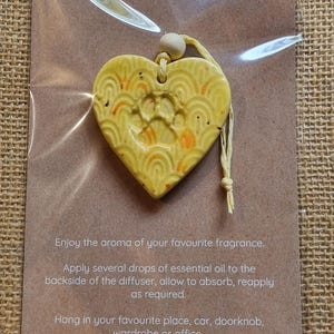 May include: A yellow ceramic heart-shaped essential oil diffuser with a paw print design. The diffuser is hanging from a string with a wooden bead. The text on the card reads: "Essential Oil Diffuser. Enjoy the aroma of your favourite fragrance. Apply several drops of essential oil to the backside of the diffuser, allow to absorb, reapply as required. Hang in your favourite place, car, doorknob, wardrobe or office."