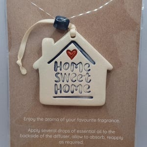 May include: A ceramic essential oil diffuser shaped like a house, inscribed with "Home Sweet Home" in blue lettering, topped with a red heart. It hangs from a cream ribbon. The words "ESSENTIAL OIL DIFFUSER" are printed at the top of the card.