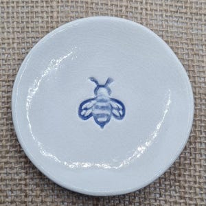 May include: A small, round, white ceramic dish featuring a blue bee motif in the centre. The dish has a textured surface and a raised edge. The bee is detailed with blue stripes and wings. Suitable for small items.