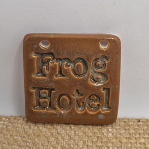 May include: A small, square, brown ceramic tile with the words "Frog Hotel" embossed in black lettering. The tile has two holes at the top for hanging.