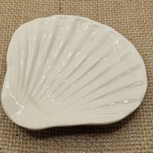 May include: Cream-coloured, shell-shaped dish with a textured surface. The dish has a scalloped edge and a fan-like pattern radiating from the centre. The dish is made of ceramic or porcelain and is likely used for decorative purposes.
