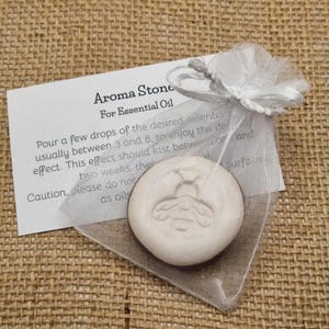 May include: Aroma stone for essential oil, presented in a sheer white bag with a matching ribbon. The stone is round, off-white, and has a bee design. A white card with instructions is included.