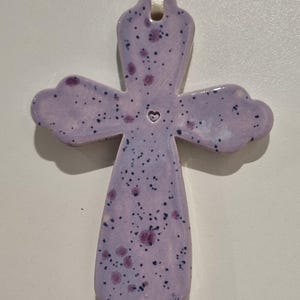 May include: A purple ceramic cross with a small heart in the centre. The cross is decorated with white and purple speckles.