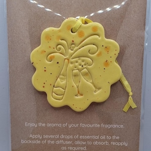 May include: Yellow ceramic essential oil diffuser with a champagne bottle and glasses design. The diffuser has a scalloped edge and orange speckles. Text at the top reads "ESSENTIAL OIL DIFFUSER". Instructions are also included.