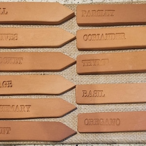 May include: Ten terracotta plant markers with different herb names, including dill, chives, rocket, sage, rosemary, mint, parsley, coriander, thyme, basil, and oregano.