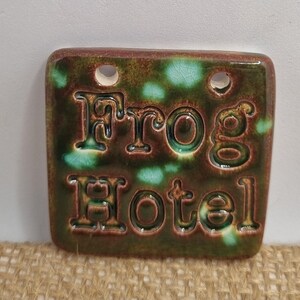 May include: A green and brown ceramic tile with the words "Frog Hotel" in raised lettering. The tile has two holes in the top for hanging.