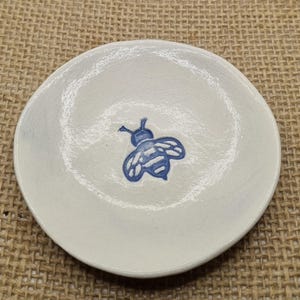 May include: A small, round, white ceramic plate with a hand-painted blue bee design in the centre. The plate has a slightly raised edge and a smooth surface. The bee is detailed with visible wings and body segments.