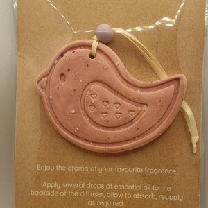 May include: A pink, bird-shaped essential oil diffuser with a speckled texture and heart details, hanging from a beige ribbon. The diffuser is displayed on a brown card with the text "ESSENTIAL OIL DIFFUSER" and instructions for use.