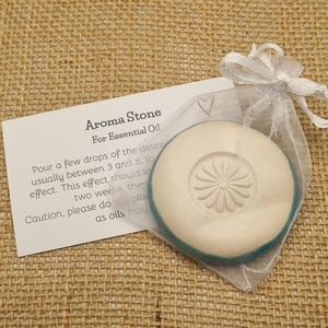 May include: Aroma Stone for essential oils. The white stone with a floral design is inside a sheer white bag tied with a ribbon. A card with instructions is next to the bag. The stone is approximately 5 cm in diameter.