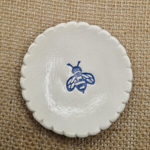 May include: A small, white ceramic dish with a scalloped edge. The dish has a blue bee design in the centre. The bee is detailed with stripes and wings. The dish is round and appears to be handmade.
