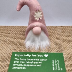 May include: A pink ceramic gnome with a white beard and a flower design on its hat. The gnome is displayed with a green card that reads "Especially for You" and promises good fortune and protection.