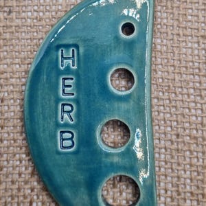 May include: A teal ceramic herb marker with the word "HERB" vertically displayed. The marker has a semi-circular shape with three circular holes and one smaller hole. The background is a textured, tan fabric.