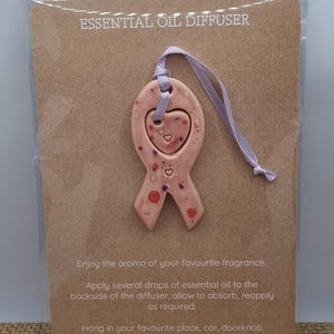 May include: A pink ceramic essential oil diffuser in a ribbon shape, with a heart-shaped cutout and small red accents. The diffuser hangs from a lilac ribbon and is displayed on a brown card with instructions.