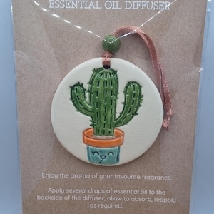 May include: A round ceramic essential oil diffuser featuring a green cactus in a blue pot with a smiling face. The diffuser is attached to a pink ribbon with a green bead and mounted on a brown cardboard backing with the text ESSENTIAL OIL DIFFUSER.