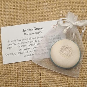 May include: Aroma stone for essential oil, in a sheer white organza bag. The round, off-white stone has a floral design. A card with instructions is included. The background is a textured, tan fabric.