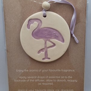 May include: A circular, cream-coloured essential oil diffuser featuring a purple flamingo. It has a purple ribbon for hanging. The text "ESSENTIAL OIL DIFFUSER" is at the top. Instructions are printed below.
