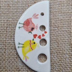 May include: A white, semi-circular ceramic decorative piece featuring two cartoon birds. One bird is pink, the other yellow, with red heart accents. The piece has three holes and is likely a decorative element.