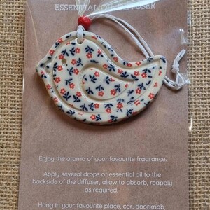 May include: A white ceramic bird-shaped diffuser with a red and blue floral pattern. The diffuser is hanging from a white string and has a red bead at the top. The text "ESSENTIAL OIL DIFFUSER" is printed on the card behind the diffuser. The card also includes instructions on how to use the diffuser.