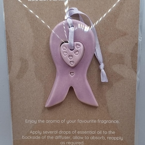 May include: A lilac-coloured essential oil diffuser in a ribbon design, featuring a heart-shaped centre with heart cutouts. It hangs from a white ribbon. The text "ESSENTIAL OIL DIFFUSER" is at the top of the card.