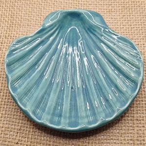 May include: A teal ceramic seashell-shaped dish. The dish has a glossy finish and detailed ridges that mimic the natural texture of a seashell. The dish is displayed on a textured, neutral-toned surface.