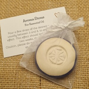 May include: Aroma Stone for essential oil, in a sheer white bag. The round, off-white stone features a floral design. A card with instructions is included. The background is a woven fabric.