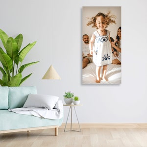 May include: A young girl in a white dress with blue embroidery jumps in the air with her arms raised. Her parents are sitting behind her on a white bed. The girl has curly brown hair and is smiling.