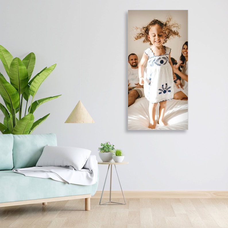 May include: A young girl in a white dress with blue embroidery jumps in the air with her arms raised. Her parents are sitting behind her on a white bed. The girl has curly brown hair and is smiling.