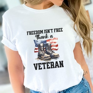 Puede incluir: Camiseta blanca con el texto "FREEDOM ISN'T FREE thank a VETERAN" y un gráfico de un par de botas marrones sobre una bandera estadounidense.