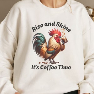 Peut inclure: Sweat-shirt blanc avec un coq tenant une tasse de café. Le texte "Rise and Shine" est au-dessus du coq et "It's Coffee Time" est en dessous du coq.