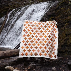 May include: A rectangular quilt featuring a repeating pattern of orange and brown cross-shaped blocks arranged on a white background. The quilt displays a grid-like design with consistent spacing between each cross, creating a geometric aesthetic.