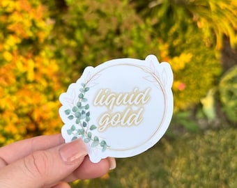May include: A white sticker featuring the words "liquid gold" in a cursive font, encircled by a gold border and green leaves. The sticker is held in a hand against a blurred backdrop of green and yellow foliage.