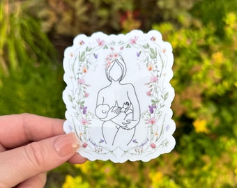 May include: A white sticker featuring a line drawing of a person breastfeeding a baby, framed by a floral wreath in pink, purple, and green. The sticker has a scalloped edge and is set against a blurred green backdrop.