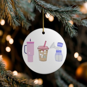 May include: A white ceramic Christmas ornament with three illustrations: a pink tumbler, an iced coffee, and a breast pump. The ornament hangs from a gold string against a blurred background of Christmas lights and pine needles.