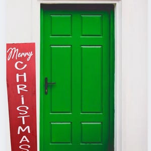 May include: A bright green door with a black handle and a red wooden sign that says "Merry Christmas" in white script. The sign is leaning against the door frame. The door is set in a white frame.