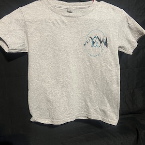 May include: A gray t-shirt with a black and blue graphic of a mountain range with the text "G.R.O.O.V.E. GINGRAS DESIGNS" in a circle.