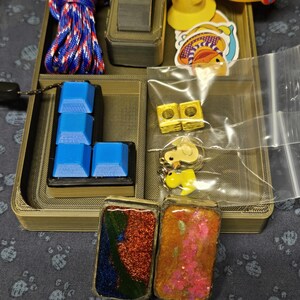 May include: A brown wooden box with various compartments. The box contains a blue and red rope, a black switch, a set of blue keyboard keys, two gold metal cubes, two yellow rubber duck toys, two small resin trinkets with glitter, and a small plastic bag with a yellow rubber duck charm.